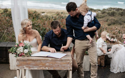 Photo shows a couple signing their marriage certificate with a coastal backdrop